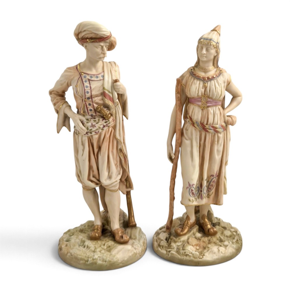 A pair of late Victorian Royal Worcester blush ivory porcelain figures depicting an Indian man and woman in detailed traditional attire with gilded accents.