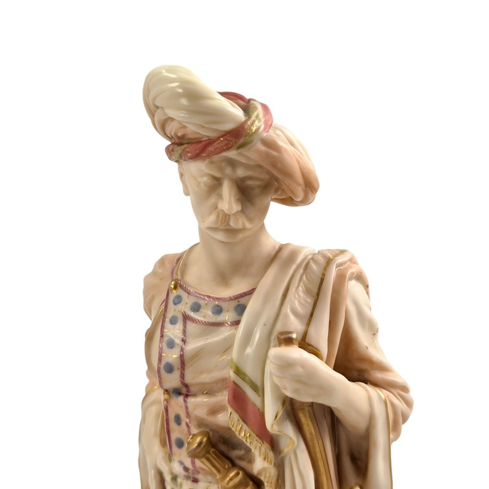 A pair of late Victorian Royal Worcester blush ivory porcelain figures depicting an Indian man and woman in detailed traditional attire with gilded accents. View X