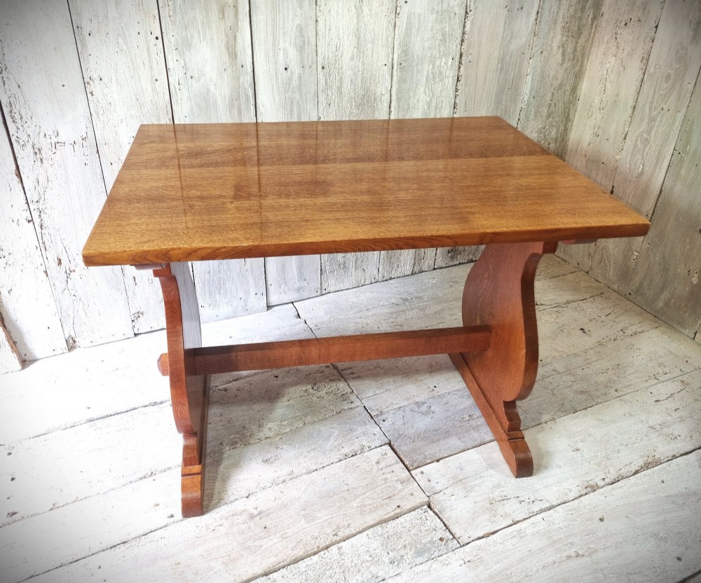A solid golden oak trestle table featuring a rectangular top, shaped side supports with visible medullary ray grain patterns, and a central stretcher secured with pegged tenons. View II