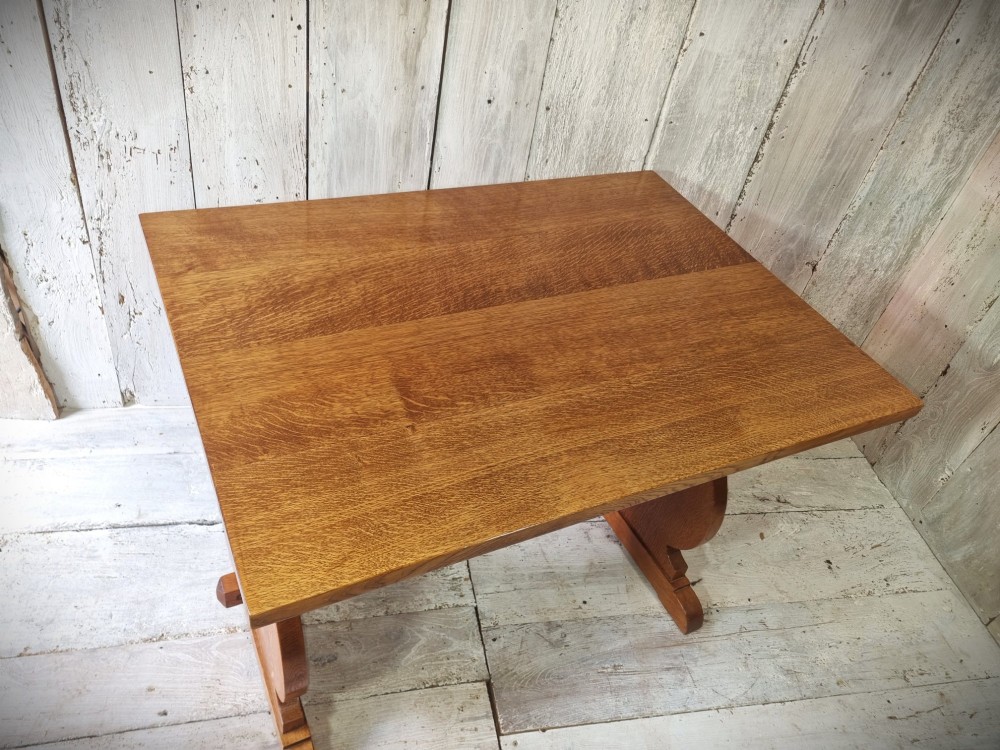 A solid golden oak trestle table featuring a rectangular top, shaped side supports with visible medullary ray grain patterns, and a central stretcher secured with pegged tenons. View III