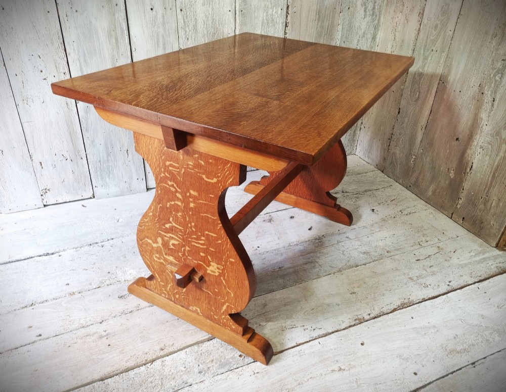 A solid golden oak trestle table featuring a rectangular top, shaped side supports with visible medullary ray grain patterns, and a central stretcher secured with pegged tenons. View IV