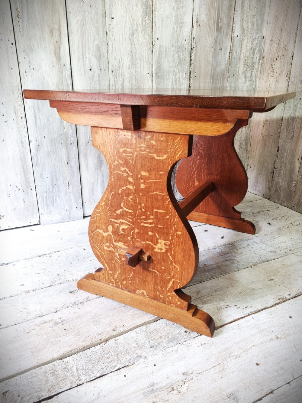 A solid golden oak trestle table featuring a rectangular top, shaped side supports with visible medullary ray grain patterns, and a central stretcher secured with pegged tenons. View V
