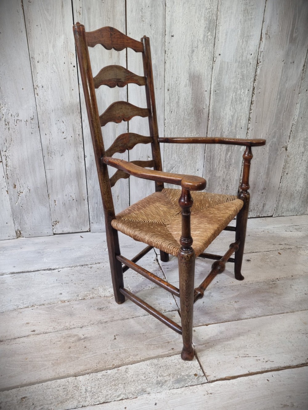 An antique Cheshire ladder back armchair featuring shaped horizontal slats, swept armrests, turned legs, and a woven rush seat. View II