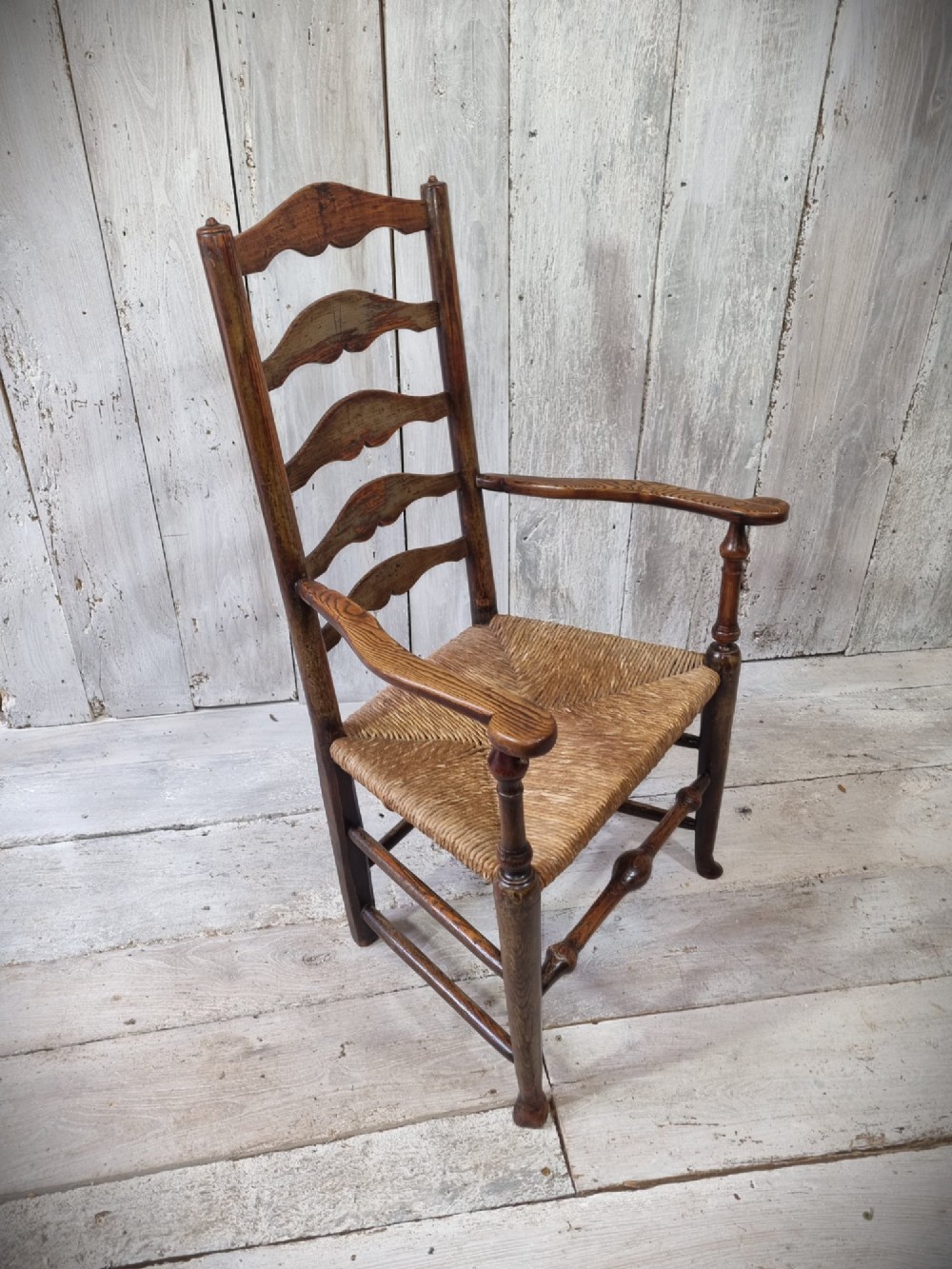 An antique Cheshire ladder back armchair featuring shaped horizontal slats, swept armrests, turned legs, and a woven rush seat. View III