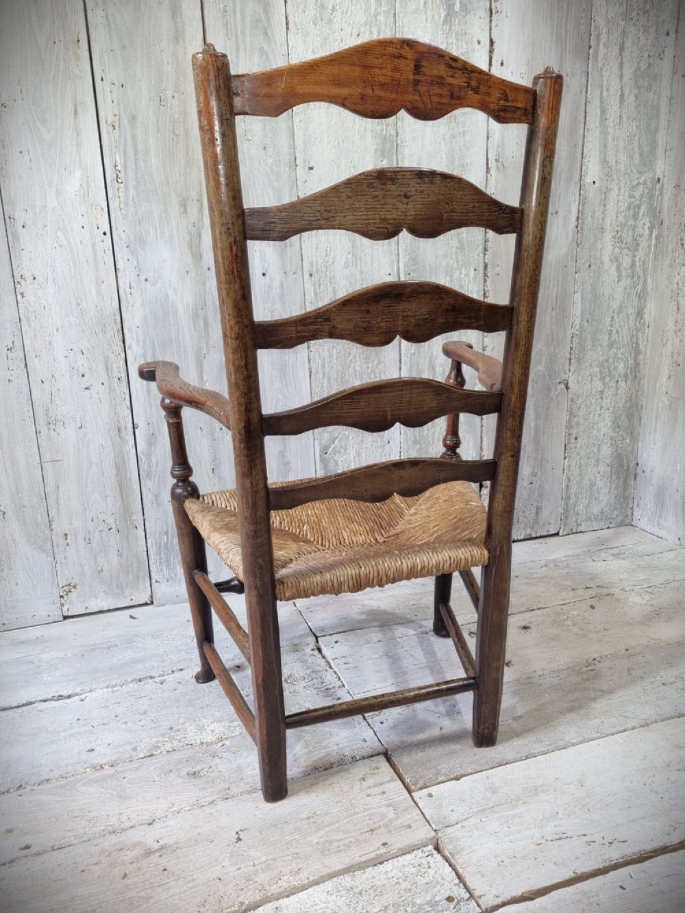 An antique Cheshire ladder back armchair featuring shaped horizontal slats, swept armrests, turned legs, and a woven rush seat. View IV