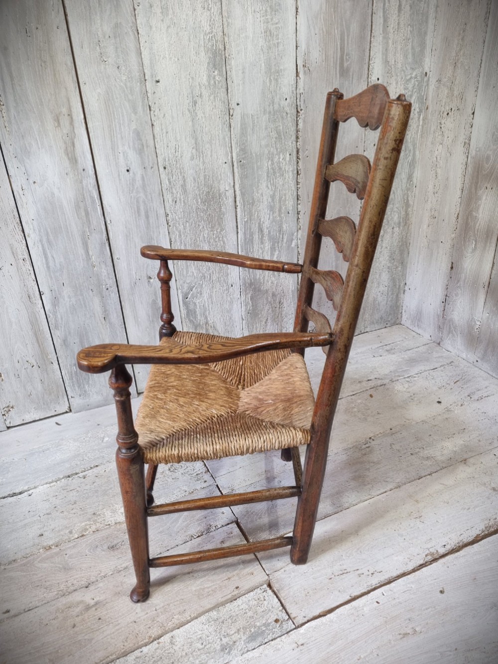 An antique Cheshire ladder back armchair featuring shaped horizontal slats, swept armrests, turned legs, and a woven rush seat. View V