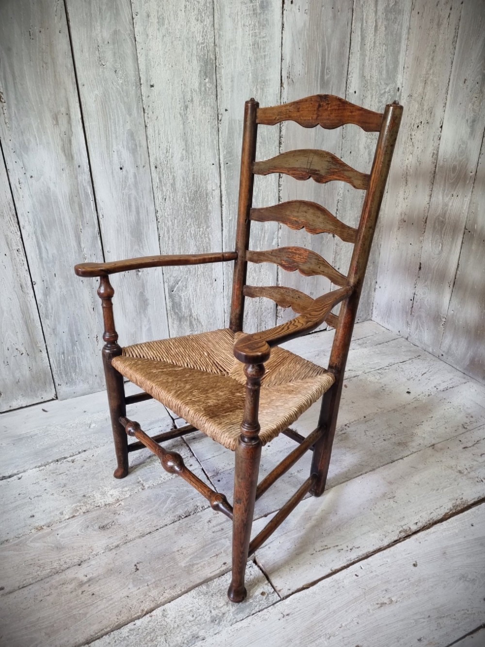 An antique Cheshire ladder back armchair featuring shaped horizontal slats, swept armrests, turned legs, and a woven rush seat. View VI