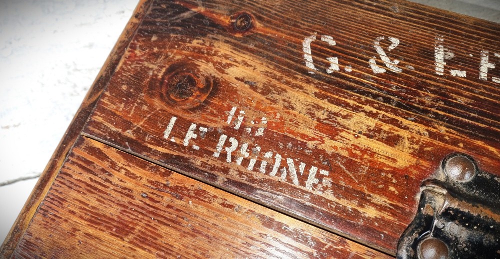 A rectangular warm brown wooden tool box featuring white stencilled text, a central winged logo, and an iron top clasp. View VI