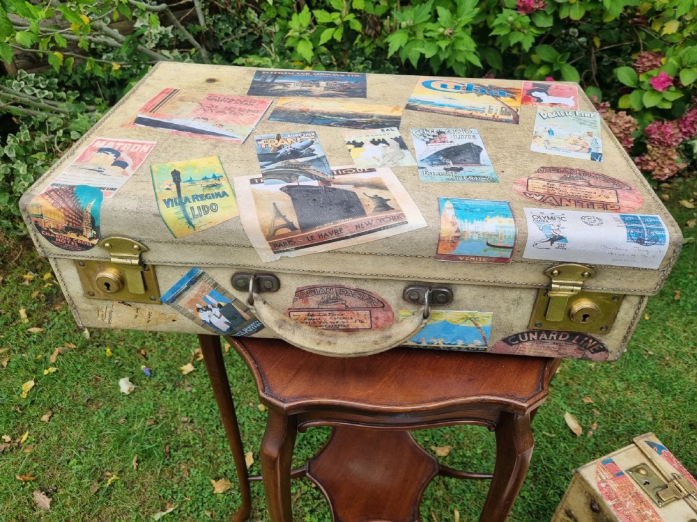 A graduated stack of three cream vellum suitcases covered in colorful travel stickers and fitted with brass latches. View V