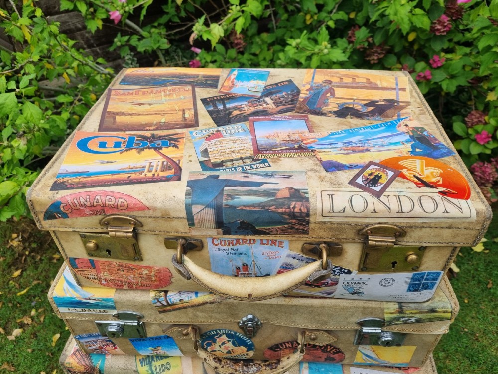 A graduated stack of three cream vellum suitcases covered in colorful travel stickers and fitted with brass latches. View VII