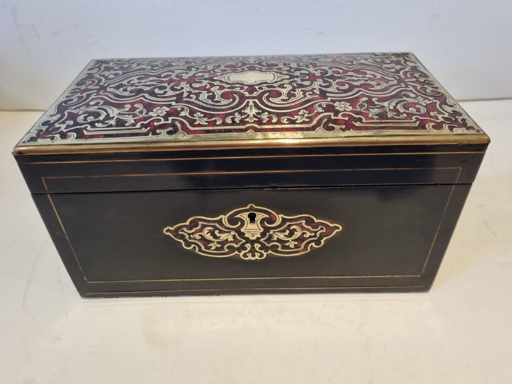 A rectangular ebonised wood tea caddy featuring a lid ornamented with red tortoiseshell and brass Boulle marquetry and a brass-inlaid keyhole escutcheon on the front.