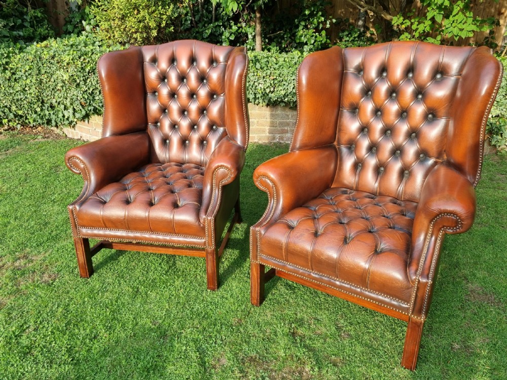 A pair of George III style chestnut brown leather Chesterfield wingback armchairs with deep buttoned upholstery, brass studding, and square wooden legs.