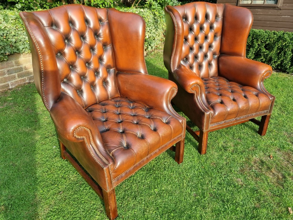 A pair of George III style chestnut brown leather Chesterfield wingback armchairs with deep buttoned upholstery, brass studding, and square wooden legs. View V