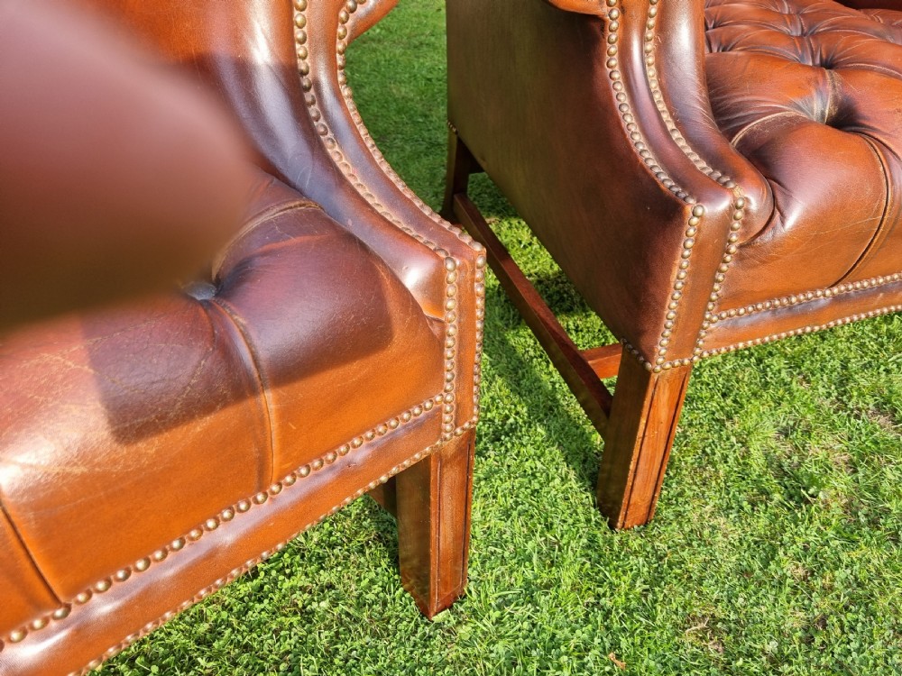 A pair of George III style chestnut brown leather Chesterfield wingback armchairs with deep buttoned upholstery, brass studding, and square wooden legs. View VI