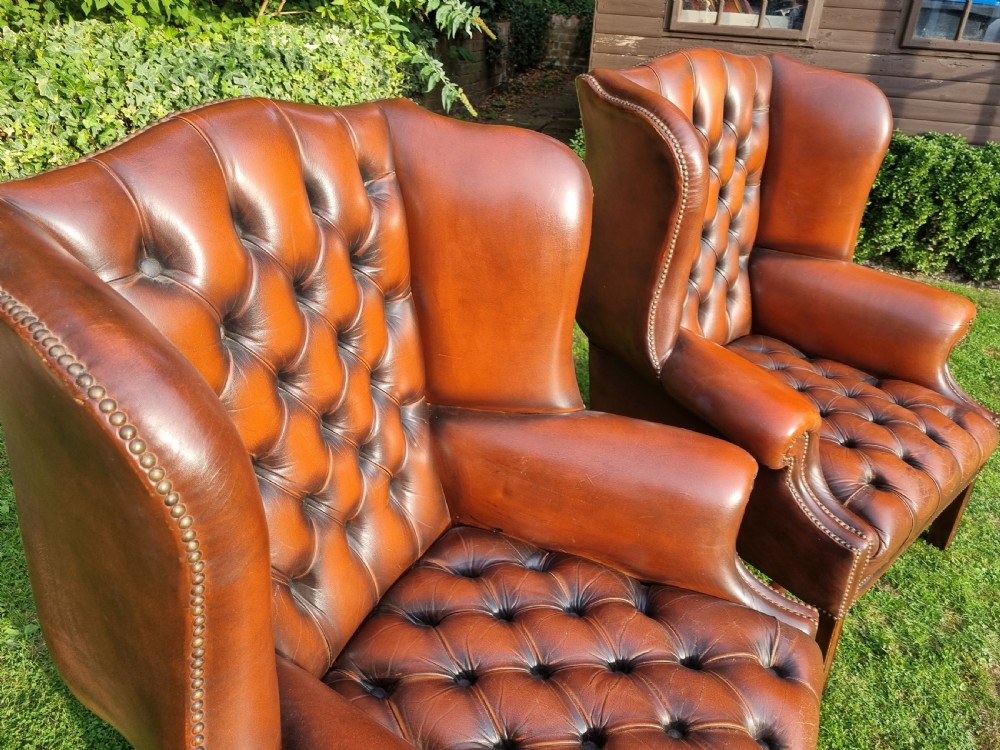 A pair of George III style chestnut brown leather Chesterfield wingback armchairs with deep buttoned upholstery, brass studding, and square wooden legs. View VII
