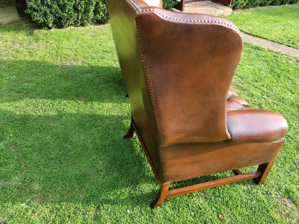 A pair of George III style chestnut brown leather Chesterfield wingback armchairs with deep buttoned upholstery, brass studding, and square wooden legs. View VIII