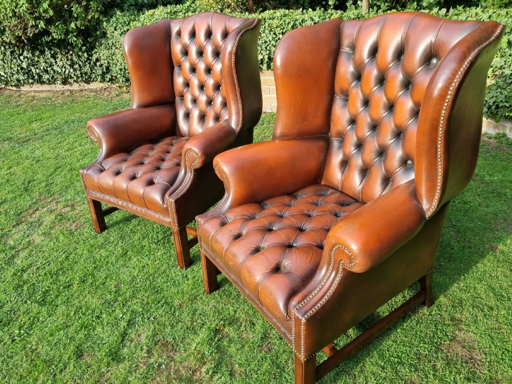 A pair of George III style chestnut brown leather Chesterfield wingback armchairs with deep buttoned upholstery, brass studding, and square wooden legs. View X