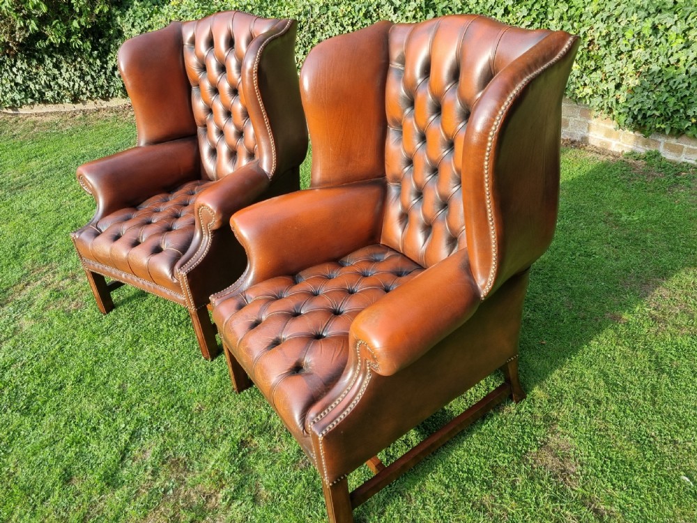 A pair of George III style chestnut brown leather Chesterfield wingback armchairs with deep buttoned upholstery, brass studding, and square wooden legs. View XII