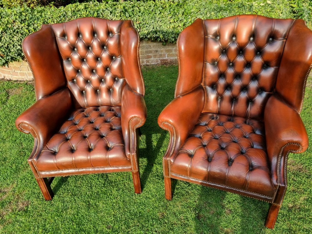 A pair of George III style chestnut brown leather Chesterfield wingback armchairs with deep buttoned upholstery, brass studding, and square wooden legs. View XIII