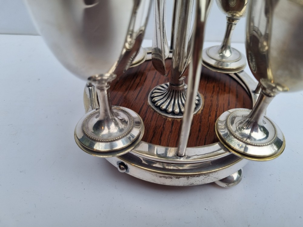 A Victorian silver-plated four-piece egg cruet featuring four goblets, four hanging spoons, a central carrying ring, and a circular oak base. View III