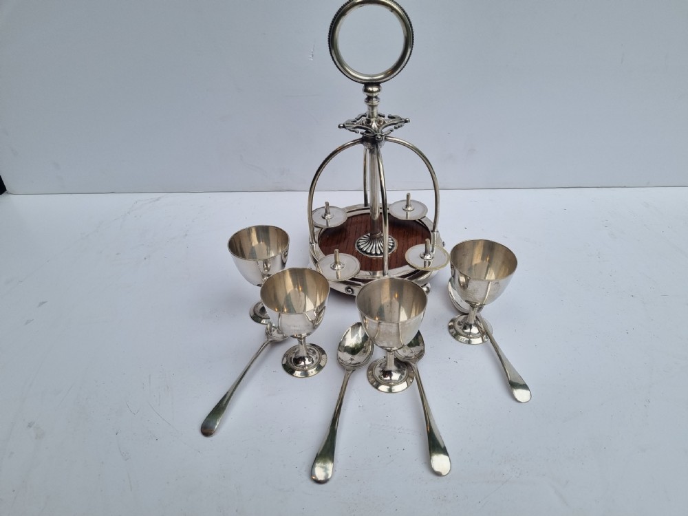 A Victorian silver-plated four-piece egg cruet featuring four goblets, four hanging spoons, a central carrying ring, and a circular oak base. View V