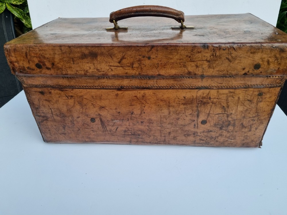 A Victorian tan leather travel trunk featuring hand-stitched edges, brass lever locks, a top carrying handle, and patinated surface wear. View VI