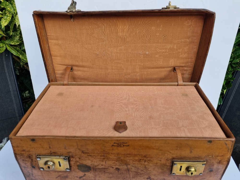 A Victorian tan leather travel trunk featuring hand-stitched edges, brass lever locks, a top carrying handle, and patinated surface wear. View X