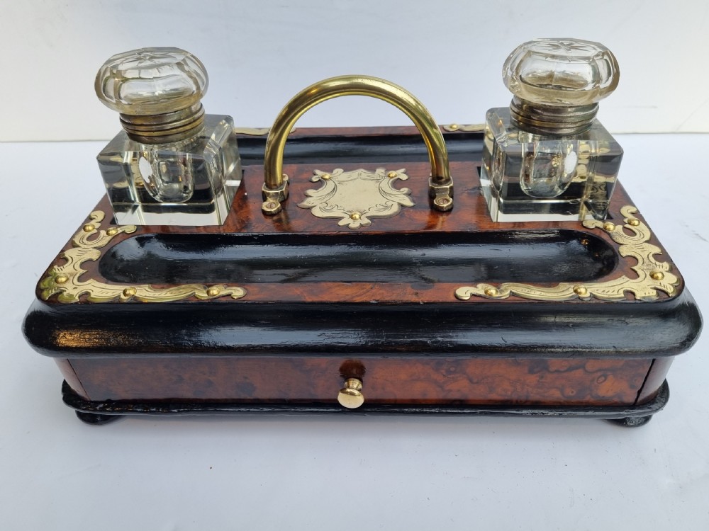 A Victorian walnut inkstand featuring two square glass inkwells, ornamental brass corner mounts, a central brass handle, a carved pen groove, and a lower drawer.