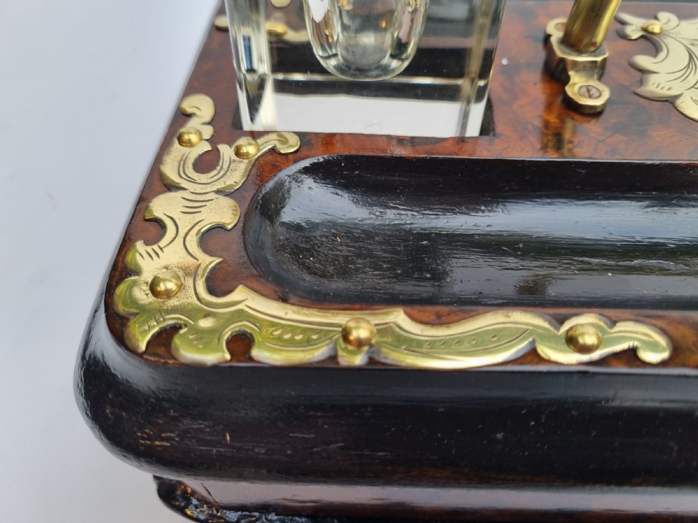 A Victorian walnut inkstand featuring two square glass inkwells, ornamental brass corner mounts, a central brass handle, a carved pen groove, and a lower drawer. View II