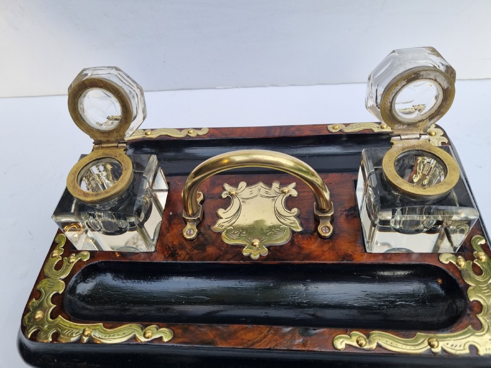 A Victorian walnut inkstand featuring two square glass inkwells, ornamental brass corner mounts, a central brass handle, a carved pen groove, and a lower drawer. View V