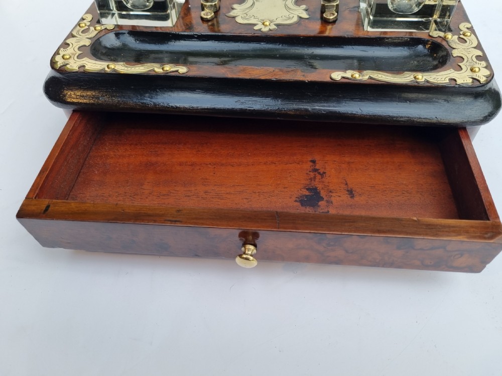 A Victorian walnut inkstand featuring two square glass inkwells, ornamental brass corner mounts, a central brass handle, a carved pen groove, and a lower drawer. View VII