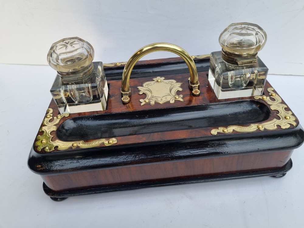 A Victorian walnut inkstand featuring two square glass inkwells, ornamental brass corner mounts, a central brass handle, a carved pen groove, and a lower drawer. View VIII