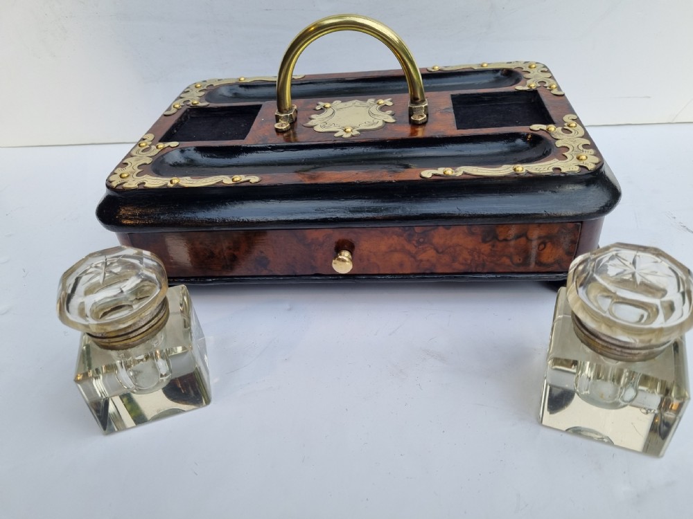 A Victorian walnut inkstand featuring two square glass inkwells, ornamental brass corner mounts, a central brass handle, a carved pen groove, and a lower drawer. View IX