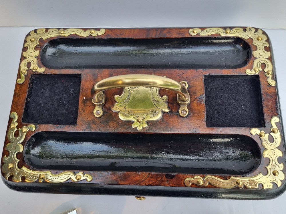 A Victorian walnut inkstand featuring two square glass inkwells, ornamental brass corner mounts, a central brass handle, a carved pen groove, and a lower drawer. View X