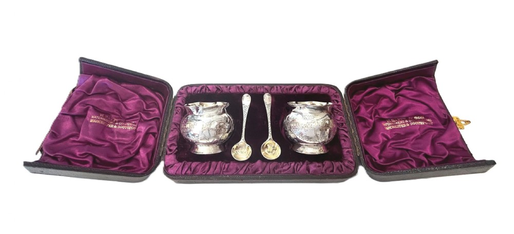 A pair of Victorian engraved sterling silver salt cellars with matching spoons displayed inside an open fitted purple silk-lined presentation case.