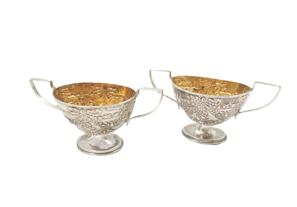 A pair of Victorian sterling silver open salt cellars featuring pedestal feet, double handles, embossed festoon decoration, and gilt interiors. View IV
