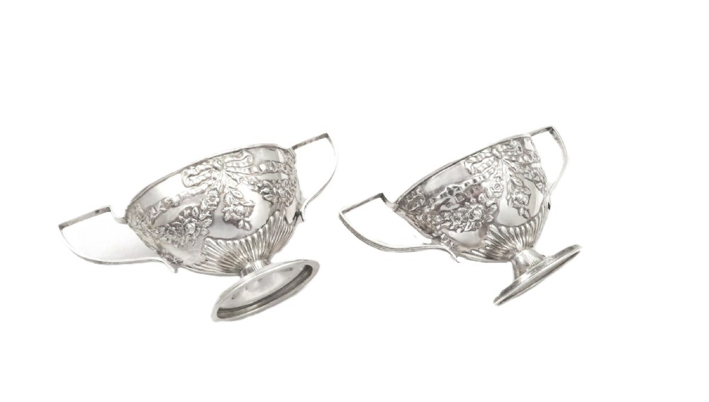 A pair of Victorian sterling silver open salt cellars featuring pedestal feet, double handles, embossed festoon decoration, and gilt interiors. View V