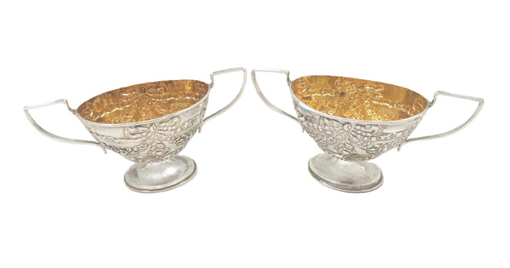 A pair of Victorian sterling silver open salt cellars featuring pedestal feet, double handles, embossed festoon decoration, and gilt interiors. View VI