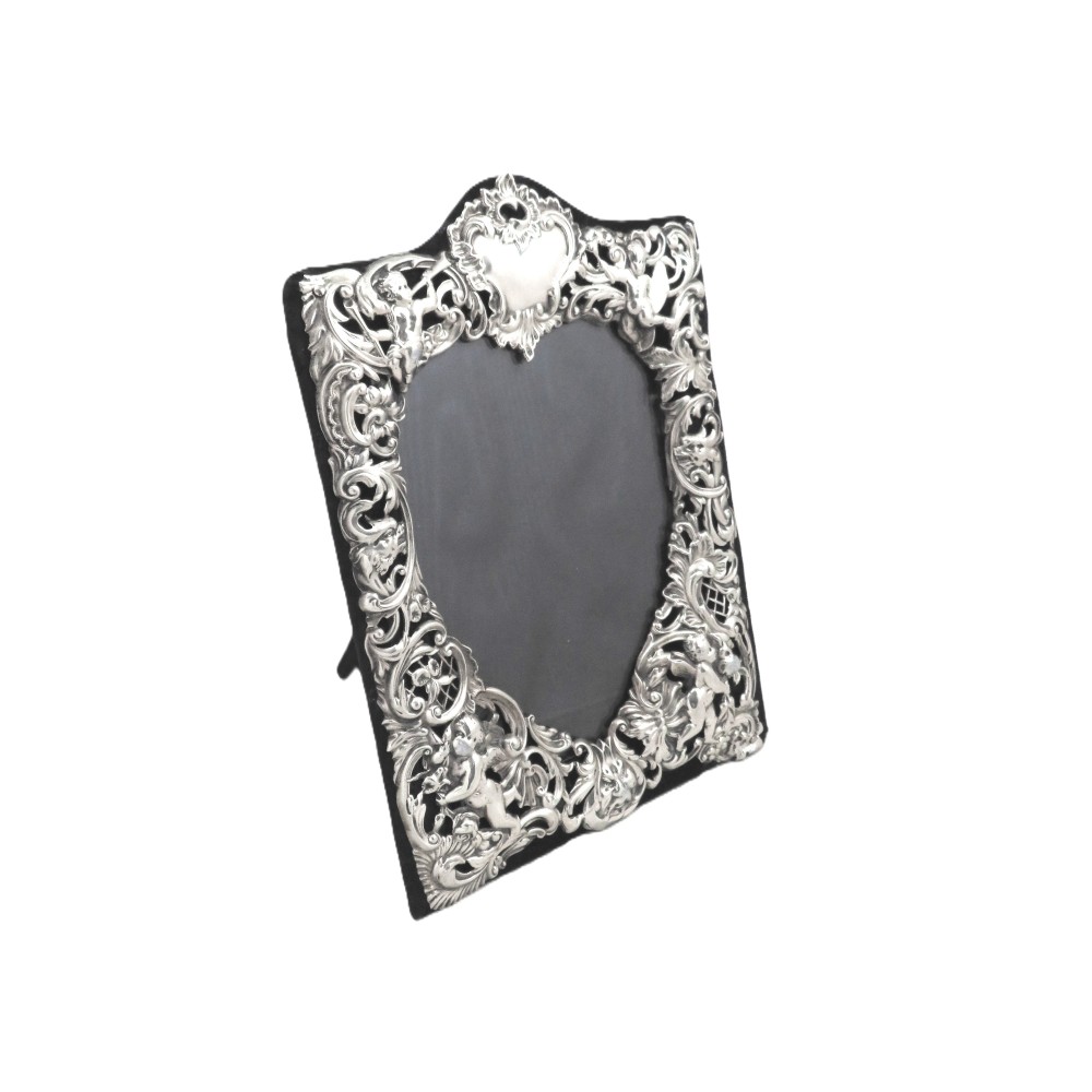 An antique Victorian sterling silver easel photo frame featuring a heart-shaped central opening surrounded by embossed cherubs, foliate scrolls, and a blank top cartouche.