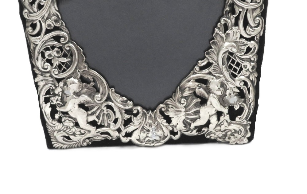 An antique Victorian sterling silver easel photo frame featuring a heart-shaped central opening surrounded by embossed cherubs, foliate scrolls, and a blank top cartouche. View VII