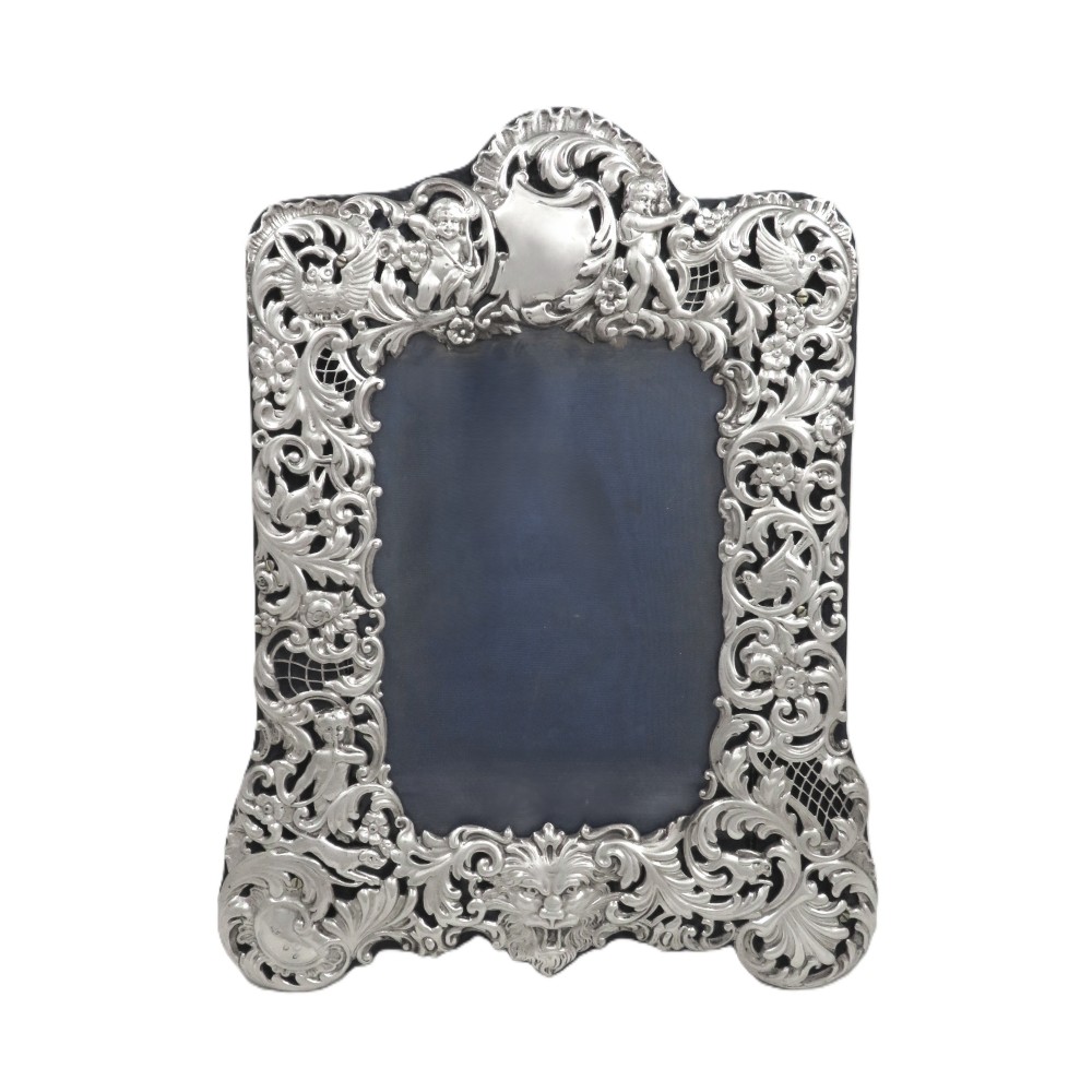 An ornate late Victorian sterling silver photo frame featuring intricate pierced scrollwork, cherub figures, birds, and a central lion mask at the base.