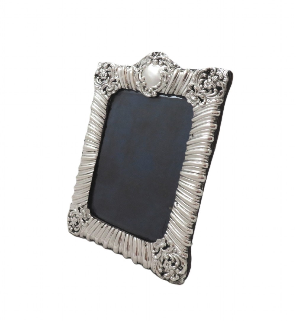 An antique Edwardian sterling silver picture frame featuring fluted borders, floral repousse details, a central top cartouche, and an easel backing.
