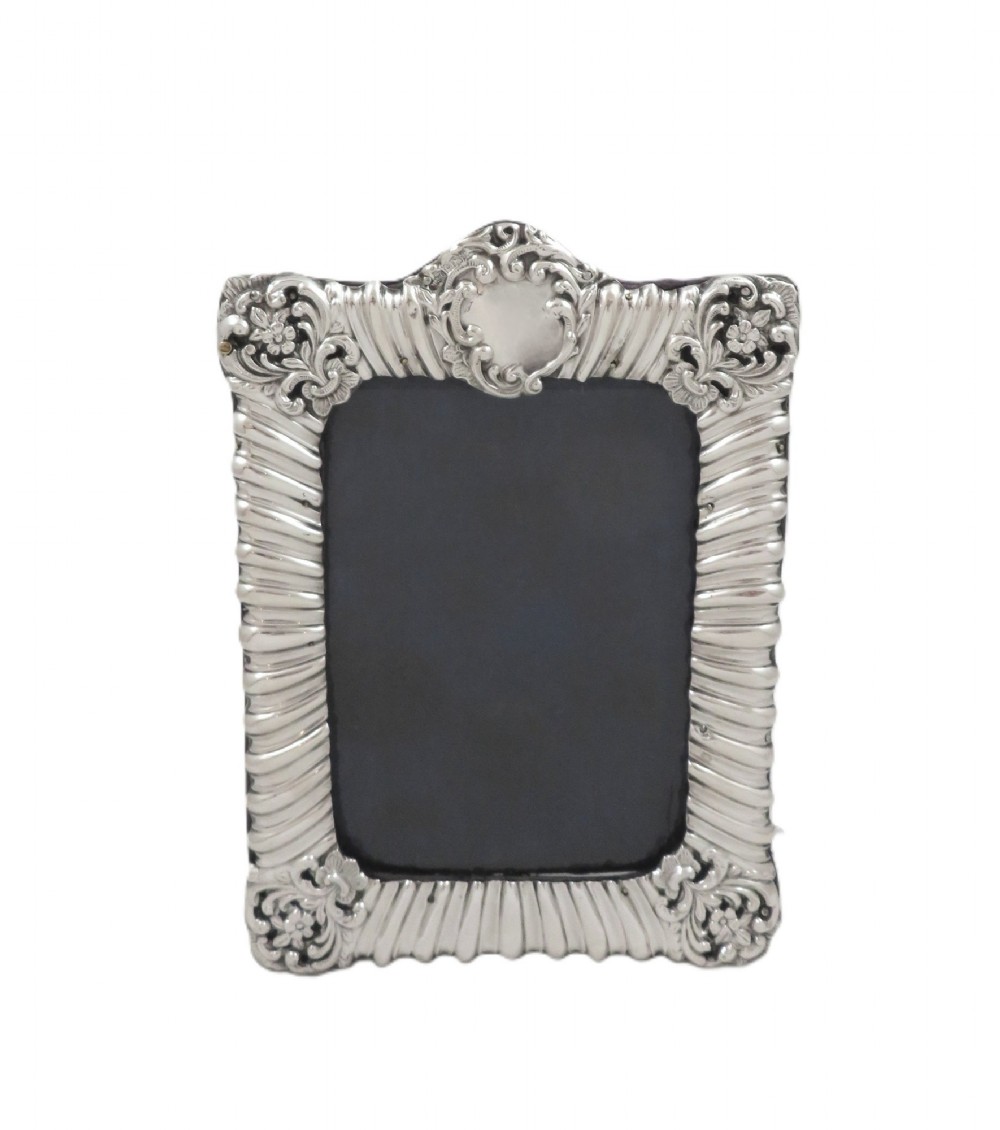 An antique Edwardian sterling silver picture frame featuring fluted borders, floral repousse details, a central top cartouche, and an easel backing. View V