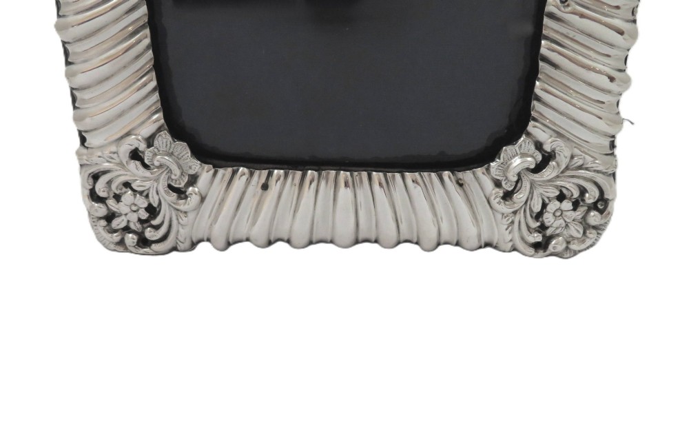 An antique Edwardian sterling silver picture frame featuring fluted borders, floral repousse details, a central top cartouche, and an easel backing. View VI