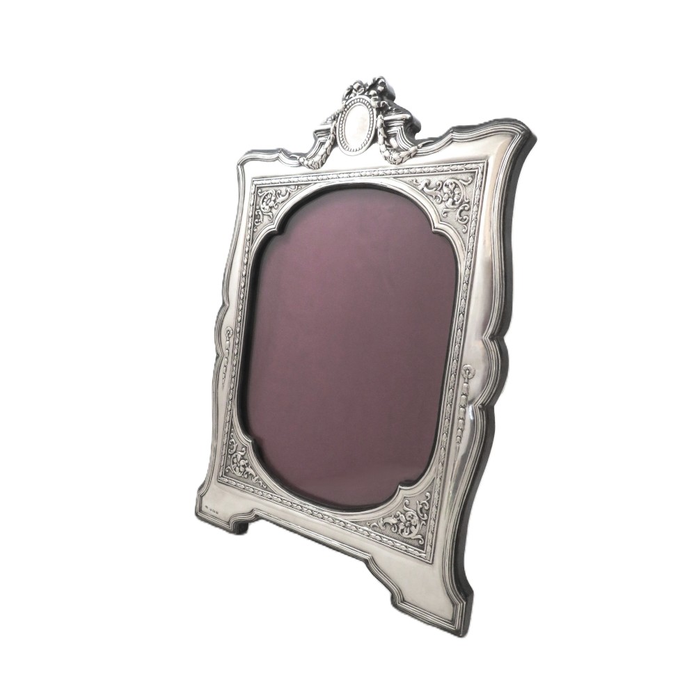 An ornate Edwardian sterling silver portrait photo frame featuring a ribbon crest and shaped border, resting on an easel back.