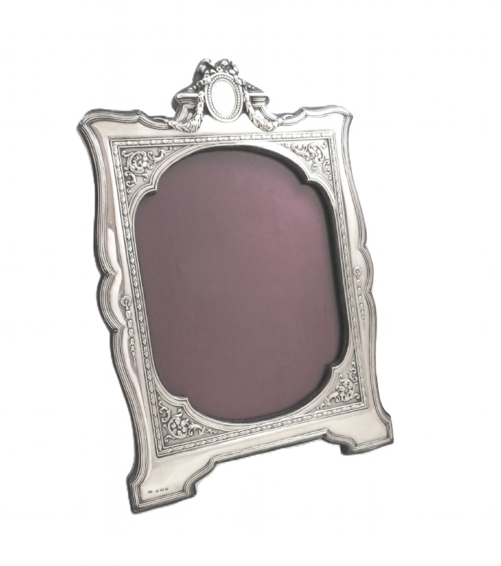 An ornate Edwardian sterling silver portrait photo frame featuring a ribbon crest and shaped border, resting on an easel back. View IV