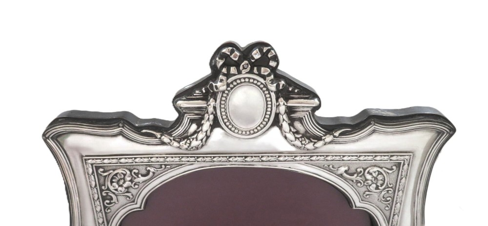 An ornate Edwardian sterling silver portrait photo frame featuring a ribbon crest and shaped border, resting on an easel back. View VI