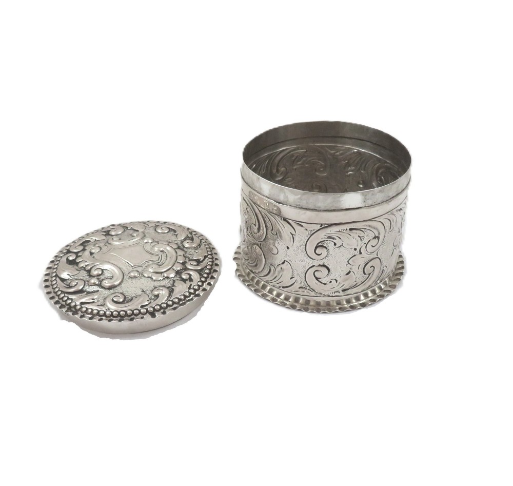A small cylindrical Victorian sterling silver vanity pot decorated with floral repousse scrollwork and a beaded rim around the lid. View V