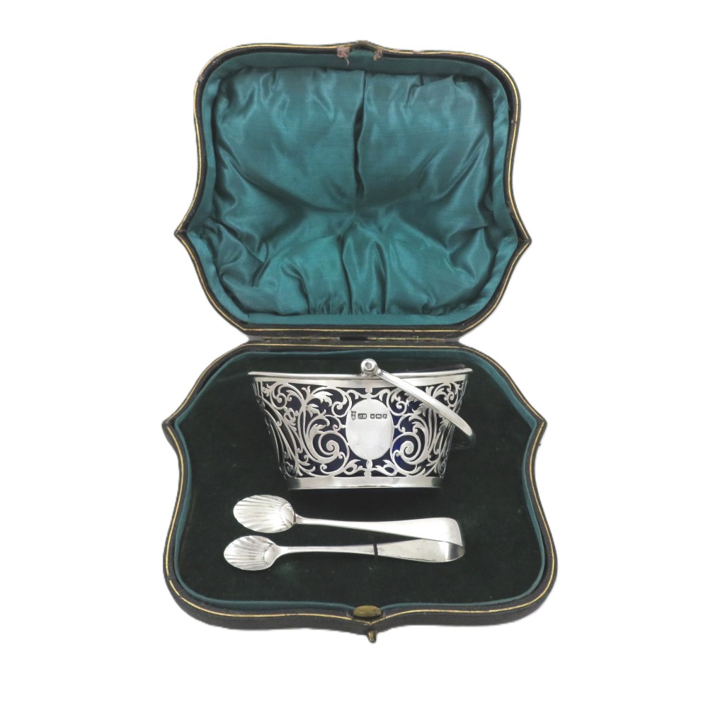 An Edwardian hallmarked sterling silver pierced sugar basket with a cobalt blue glass liner and matching silver sugar tongs in an open presentation case.
