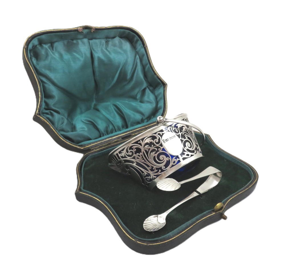 An Edwardian hallmarked sterling silver pierced sugar basket with a cobalt blue glass liner and matching silver sugar tongs in an open presentation case. View IV